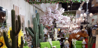 A vibrant Mexican-themed home decor display featuring large artificial cacti, bright green patio furniture, sunflowers, a cow statue, and various rustic lanterns and lighting fixtures, suggesting an eclectic outdoor living space.