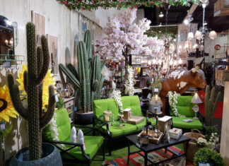 A vibrant Mexican-themed home decor display featuring large artificial cacti, bright green patio furniture, sunflowers, a cow statue, and various rustic lanterns and lighting fixtures, suggesting an eclectic outdoor living space.