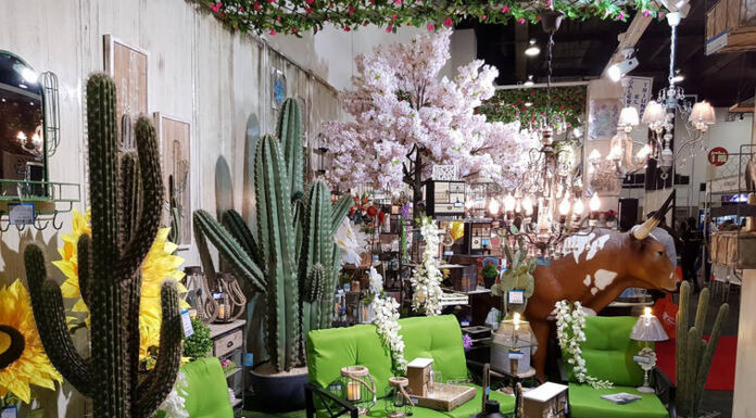 A vibrant Mexican-themed home decor display featuring large artificial cacti, bright green patio furniture, sunflowers, a cow statue, and various rustic lanterns and lighting fixtures, suggesting an eclectic outdoor living space.