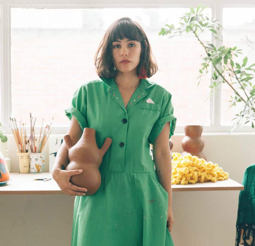 A Mexican female artist with a bob haircut and a green dress holds a unique, organically shaped terracotta sculpture in her studio, with art supplies and other creative elements visible in the background.