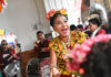 A joyous woman with flowers in her hair, wearing traditional Mexican attire and a necklace with large medallions, sings and dances while a band plays in the background.