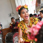 A joyous woman with flowers in her hair, wearing traditional Mexican attire and a necklace with large medallions, sings and dances while a band plays in the background.