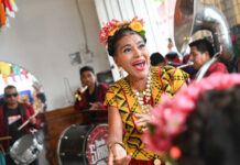 A joyous woman with flowers in her hair, wearing traditional Mexican attire and a necklace with large medallions, sings and dances while a band plays in the background.