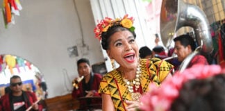 A joyous woman with flowers in her hair, wearing traditional Mexican attire and a necklace with large medallions, sings and dances while a band plays in the background.