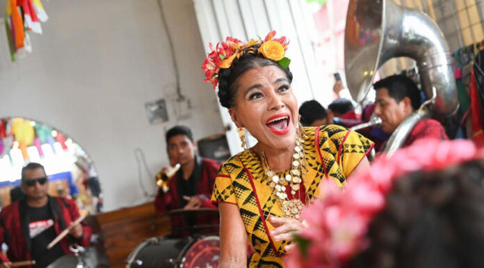A joyous woman with flowers in her hair, wearing traditional Mexican attire and a necklace with large medallions, sings and dances while a band plays in the background.