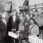 Carlos Torres standing to the left of two men in front of a chess board.