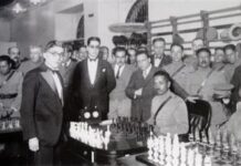 Carlos Torres standing to the left of two men in front of a chess board.