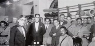 Carlos Torres standing to the left of two men in front of a chess board.