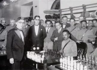 Carlos Torres standing to the left of two men in front of a chess board.