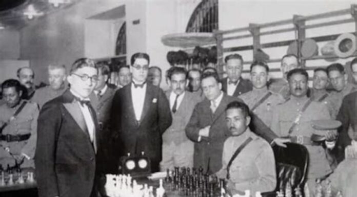 Carlos Torres standing to the left of two men in front of a chess board.