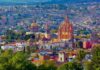 An aerial view of San Miguel de Allende, Mexico, showcasing its dense urban landscape and iconic architectural landmarks under a bright, clear sky. In the foreground, vibrant green trees partially obscure the view. The colonial-era city is characterized by warm, earthy tones like ochre, terracotta, and cream. Prominently featured in the midground is the Parroquia de San Miguel Arcángel, a striking neo-Gothic church with a pinkish-orange facade and towering spires.