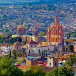 An aerial view of San Miguel de Allende, Mexico, showcasing its dense urban landscape and iconic architectural landmarks under a bright, clear sky. In the foreground, vibrant green trees partially obscure the view. The colonial-era city is characterized by warm, earthy tones like ochre, terracotta, and cream. Prominently featured in the midground is the Parroquia de San Miguel Arcángel, a striking neo-Gothic church with a pinkish-orange facade and towering spires.
