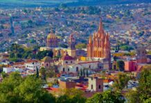 An aerial view of San Miguel de Allende, Mexico, showcasing its dense urban landscape and iconic architectural landmarks under a bright, clear sky. In the foreground, vibrant green trees partially obscure the view. The colonial-era city is characterized by warm, earthy tones like ochre, terracotta, and cream. Prominently featured in the midground is the Parroquia de San Miguel Arcángel, a striking neo-Gothic church with a pinkish-orange facade and towering spires.
