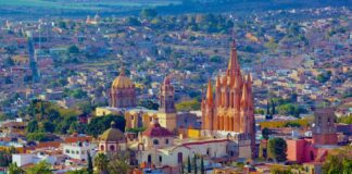An aerial view of San Miguel de Allende, Mexico, showcasing its dense urban landscape and iconic architectural landmarks under a bright, clear sky. In the foreground, vibrant green trees partially obscure the view. The colonial-era city is characterized by warm, earthy tones like ochre, terracotta, and cream. Prominently featured in the midground is the Parroquia de San Miguel Arcángel, a striking neo-Gothic church with a pinkish-orange facade and towering spires.