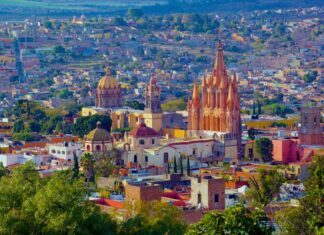 An aerial view of San Miguel de Allende, Mexico, showcasing its dense urban landscape and iconic architectural landmarks under a bright, clear sky. In the foreground, vibrant green trees partially obscure the view. The colonial-era city is characterized by warm, earthy tones like ochre, terracotta, and cream. Prominently featured in the midground is the Parroquia de San Miguel Arcángel, a striking neo-Gothic church with a pinkish-orange facade and towering spires.
