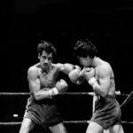 A black-and-white action shot of two boxers exchanging punches in a boxing ring. The boxer on the left, with a mustache and dark, messy hair, is throwing a left hook, while the boxer on the right is in a defensive or counterpunching stance. Both are wearing boxing gloves and shorts.