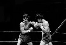 A black-and-white action shot of two boxers exchanging punches in a boxing ring. The boxer on the left, with a mustache and dark, messy hair, is throwing a left hook, while the boxer on the right is in a defensive or counterpunching stance. Both are wearing boxing gloves and shorts.