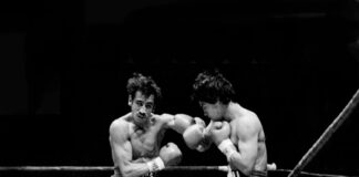 A black-and-white action shot of two boxers exchanging punches in a boxing ring. The boxer on the left, with a mustache and dark, messy hair, is throwing a left hook, while the boxer on the right is in a defensive or counterpunching stance. Both are wearing boxing gloves and shorts.