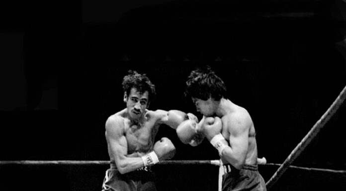 A black-and-white action shot of two boxers exchanging punches in a boxing ring. The boxer on the left, with a mustache and dark, messy hair, is throwing a left hook, while the boxer on the right is in a defensive or counterpunching stance. Both are wearing boxing gloves and shorts.