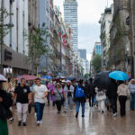 people walk through mexico city with umbrellas, with the latin america tower in the backgound