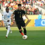 A soccer player in the black Mexican uniform dribbles ahead of another player in a white and blue uniform
