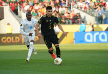 A soccer player in the black Mexican uniform dribbles ahead of another player in a white and blue uniform