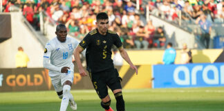 A soccer player in the black Mexican uniform dribbles ahead of another player in a white and blue uniform