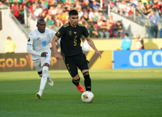 A soccer player in the black Mexican uniform dribbles ahead of another player in a white and blue uniform