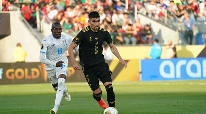 A soccer player in the black Mexican uniform dribbles ahead of another player in a white and blue uniform