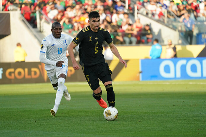 A soccer player in the black Mexican uniform dribbles ahead of another player in a white and blue uniform
