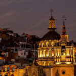A vibrant night view of Guanajuato, Mexico, featuring a grand, lit-up colonial church with a prominent dome and two spires in the foreground, set against a backdrop of tightly packed, multi-colored houses climbing the hills under a twilight sky.