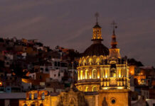 A vibrant night view of Guanajuato, Mexico, featuring a grand, lit-up colonial church with a prominent dome and two spires in the foreground, set against a backdrop of tightly packed, multi-colored houses climbing the hills under a twilight sky.