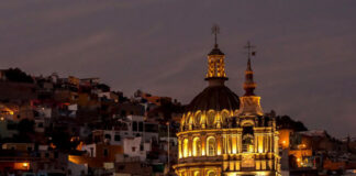 A vibrant night view of Guanajuato, Mexico, featuring a grand, lit-up colonial church with a prominent dome and two spires in the foreground, set against a backdrop of tightly packed, multi-colored houses climbing the hills under a twilight sky.
