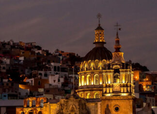 A vibrant night view of Guanajuato, Mexico, featuring a grand, lit-up colonial church with a prominent dome and two spires in the foreground, set against a backdrop of tightly packed, multi-colored houses climbing the hills under a twilight sky.