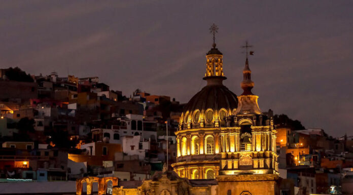 A vibrant night view of Guanajuato, Mexico, featuring a grand, lit-up colonial church with a prominent dome and two spires in the foreground, set against a backdrop of tightly packed, multi-colored houses climbing the hills under a twilight sky.