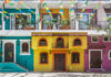 A street scene in one of Puerto Vallarta's old neighborhoods showcasing the colorful facades of multistory buildings in yellow, purple and bright teal with decorative ironwork on balconies. Overhead, strings of colorful papel picado banners are strung across the street. Potted plants adorn some windows and balconies.