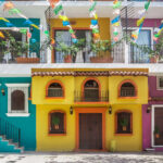 A street scene in one of Puerto Vallarta's old neighborhoods showcasing the colorful facades of multistory buildings in yellow, purple and bright teal with decorative ironwork on balconies. Overhead, strings of colorful papel picado banners are strung across the street. Potted plants adorn some windows and balconies.