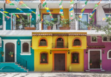 A street scene in one of Puerto Vallarta's old neighborhoods showcasing the colorful facades of multistory buildings in yellow, purple and bright teal with decorative ironwork on balconies. Overhead, strings of colorful papel picado banners are strung across the street. Potted plants adorn some windows and balconies.