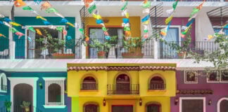 A street scene in one of Puerto Vallarta's old neighborhoods showcasing the colorful facades of multistory buildings in yellow, purple and bright teal with decorative ironwork on balconies. Overhead, strings of colorful papel picado banners are strung across the street. Potted plants adorn some windows and balconies.