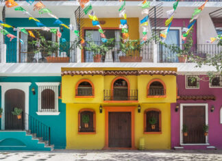 A street scene in one of Puerto Vallarta's old neighborhoods showcasing the colorful facades of multistory buildings in yellow, purple and bright teal with decorative ironwork on balconies. Overhead, strings of colorful papel picado banners are strung across the street. Potted plants adorn some windows and balconies.