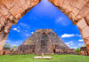 A view through a stone archway revealing the ancient, towering Pyramid of the Magician at Uxmal, a Mayan archaeological site, under a blue sky with scattered white clouds. The pyramid is surrounded by green grass and other stone ruins.