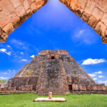 A view through a stone archway revealing the ancient, towering Pyramid of the Magician at Uxmal, a Mayan archaeological site, under a blue sky with scattered white clouds. The pyramid is surrounded by green grass and other stone ruins.