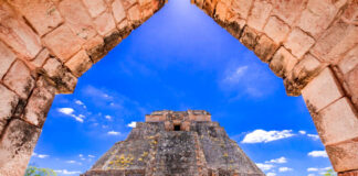 A view through a stone archway revealing the ancient, towering Pyramid of the Magician at Uxmal, a Mayan archaeological site, under a blue sky with scattered white clouds. The pyramid is surrounded by green grass and other stone ruins.