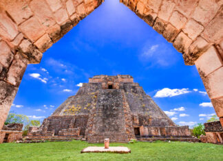 A view through a stone archway revealing the ancient, towering Pyramid of the Magician at Uxmal, a Mayan archaeological site, under a blue sky with scattered white clouds. The pyramid is surrounded by green grass and other stone ruins.