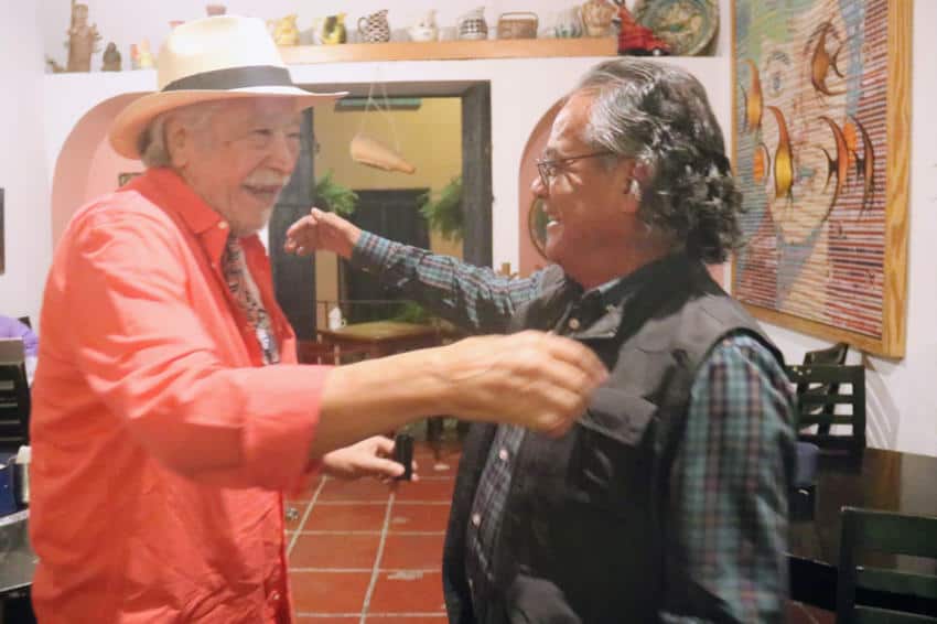 Two older Mexican mengreet each other warmly with smiles and open arms inside a home or studio filled with art and pottery.