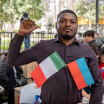 A Haitian man stands outdoors in a park-like setting, holding two small flags — one of Mexico and one of Haiti — attached to his shirt. He is looking slightly to the right with a neutral expression.