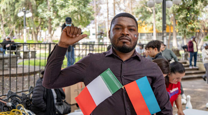 A Haitian man stands outdoors in a park-like setting, holding two small flags — one of Mexico and one of Haiti — attached to his shirt. He is looking slightly to the right with a neutral expression.