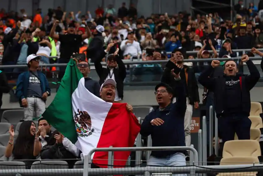 Mexican sports fans cheer passionately from the stands at a NASCAR event in Mexico. One fan man holds up a full-sized Mexican flag as he shouts in excitement.