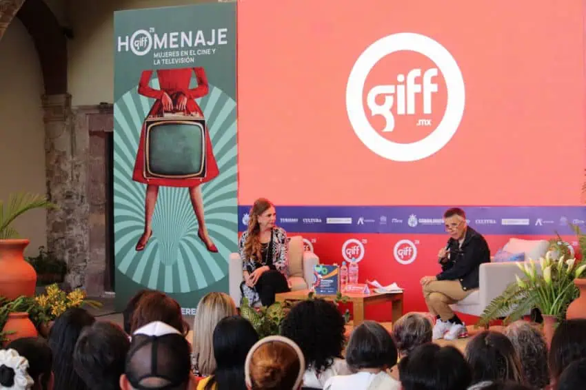 An onstage panel discussion taking place before an audience at the Guanajuato International Film Festival (GIFF) in San Miguel de Allende, Mexico. In the background, a large screen displays the festival's logo and a banner for its special tribute, "Homenaje: Mujeres en el Cine y la Televisión"