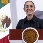 President Sheinbaum smiles from behind a podium next to a Mexican flag